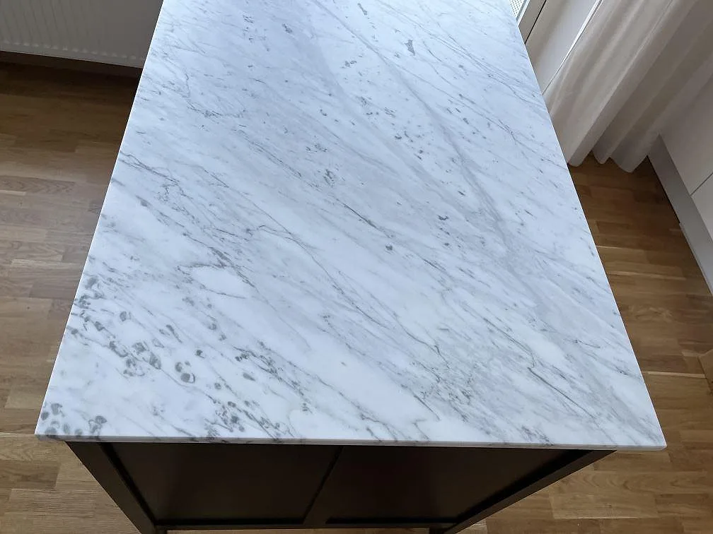 Köksö med bänkskiva i vit Carrara C marmor, matt yta med grå ådring, monterad på mörka stommar i ett kök i Göteborg.