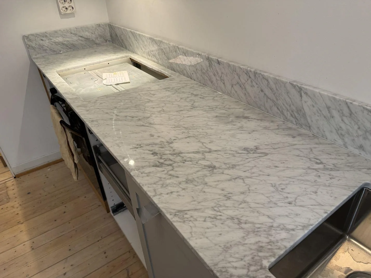 L-formad köksbänkskiva i polerad Carrara C marmor med uppvikt stänkskydd, hällurtag och rostfri diskho i ett kök i Göteborg.