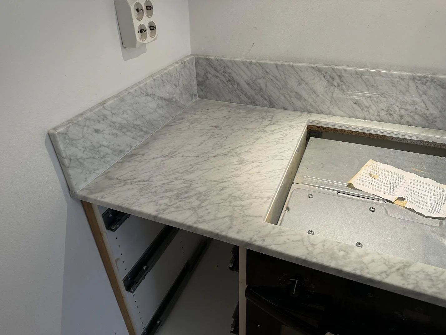 Hörn av L-formad köksbänkskiva i polerad Carrara C marmor med stänkskydd och hällurtag under montering i Göteborg.