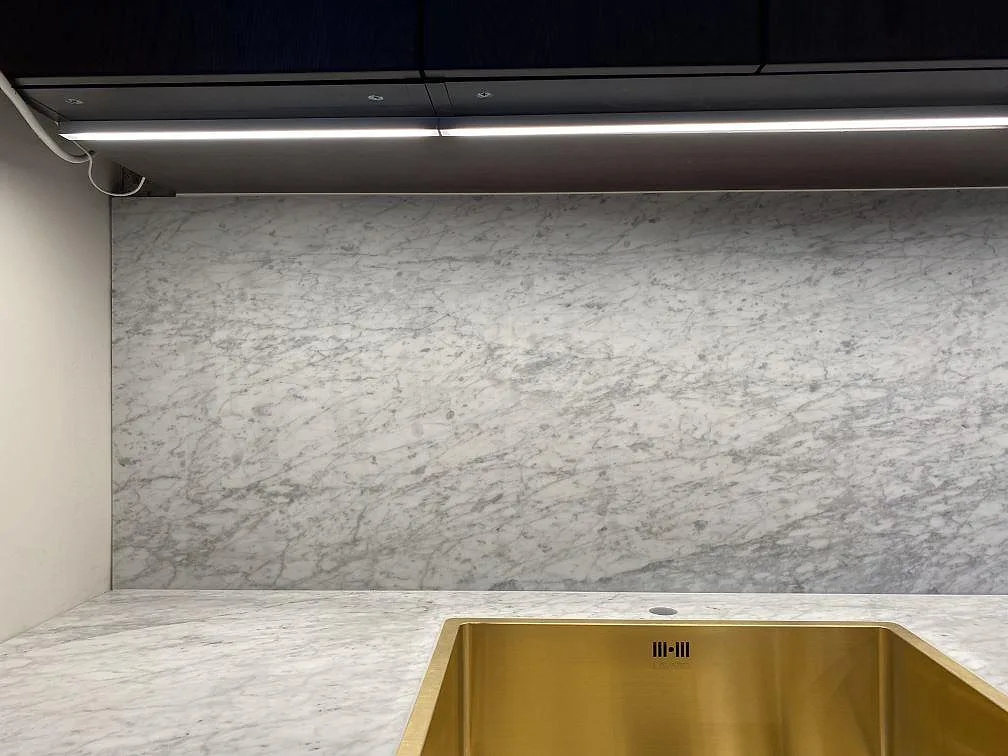 Stänkskydd och bänkskiva i matt Carrara C-marmor bakom guldfärgad diskho i Göteborg.
