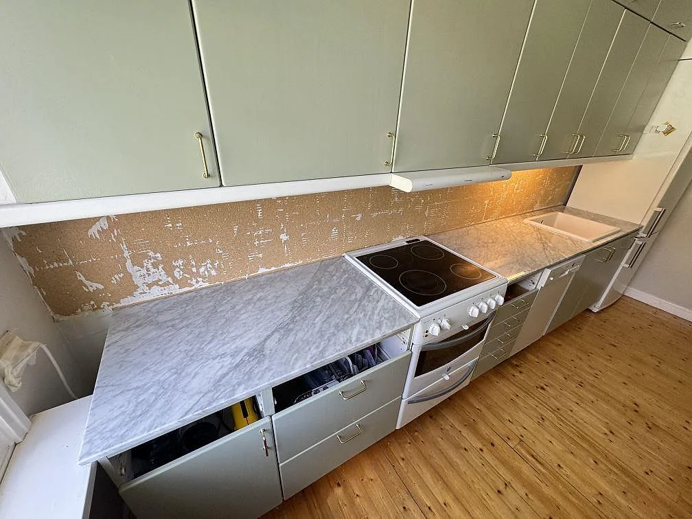 Köksbänkskiva i polerad Carrara C marmor längs en rak köksrad med spis, grågröna luckor och öppen vägg i Göteborg.