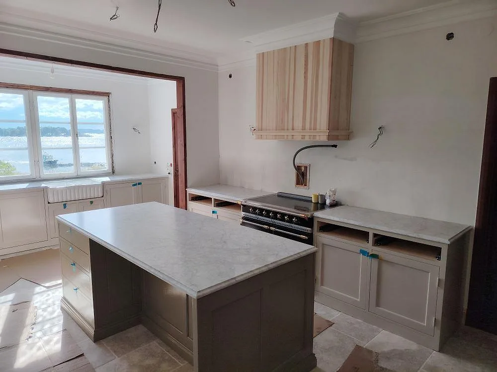 Köksbänkskiva och köksö i 30 mm matt Carrara C marmor i ett kök i Gränö med ljus träinredning, fristående spis och fönster mot vattnet.