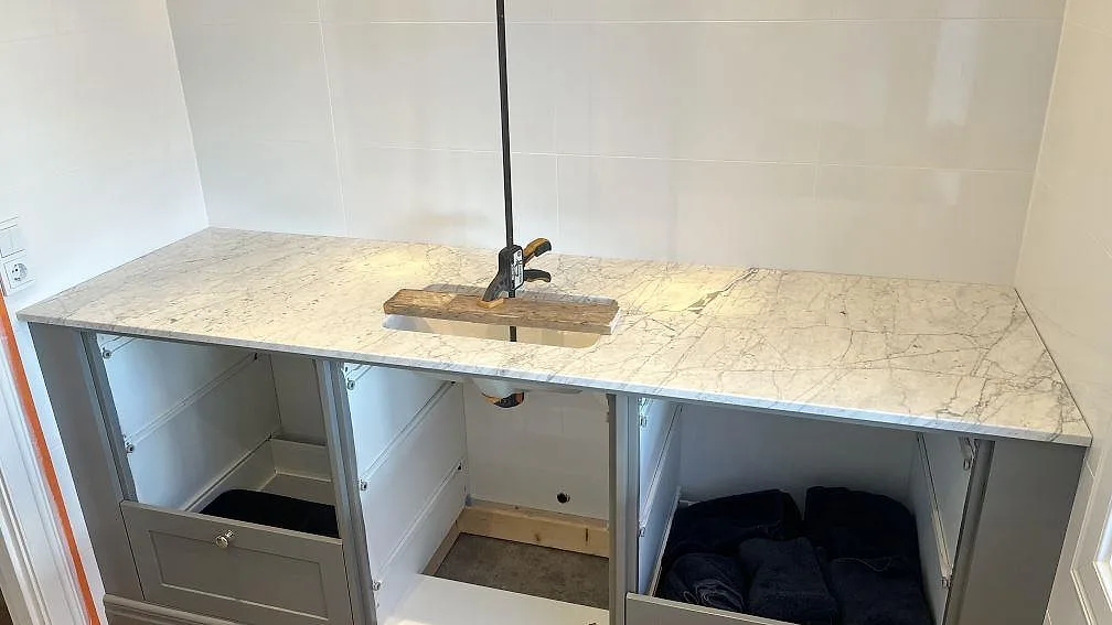 Polerad 20 mm badrumsbänkskiva i Carrara C marmor över öppna grå stommar i ett badrum i Granbersdal.