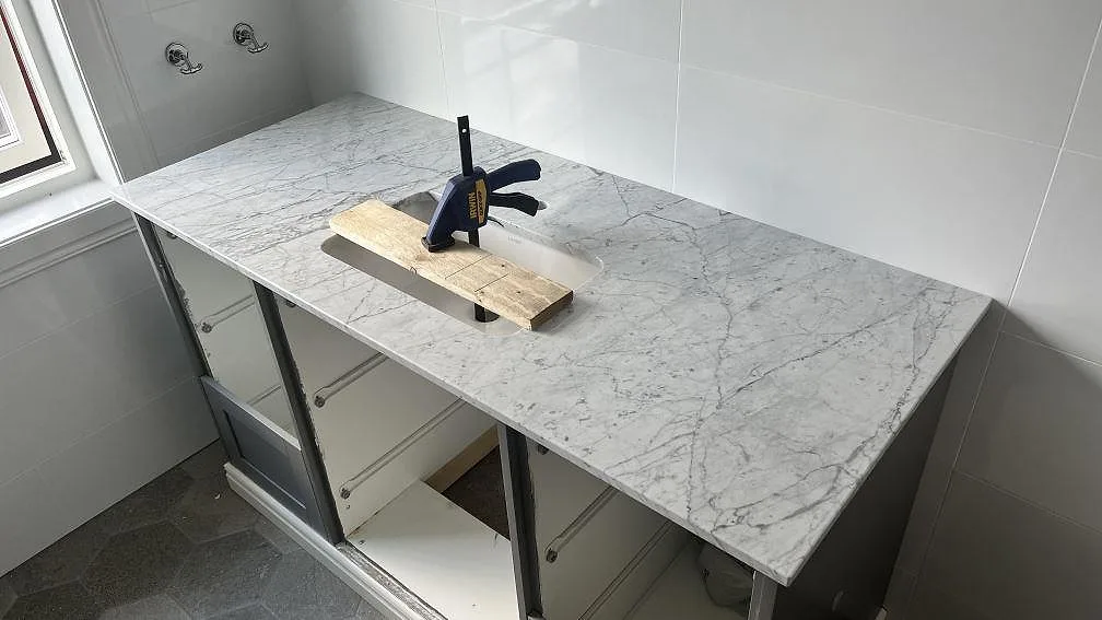 Närbild av polerad Carrara C marmor som badrumsbänkskiva med synlig grå ådring och vägganslutningar i Granbersdal.
