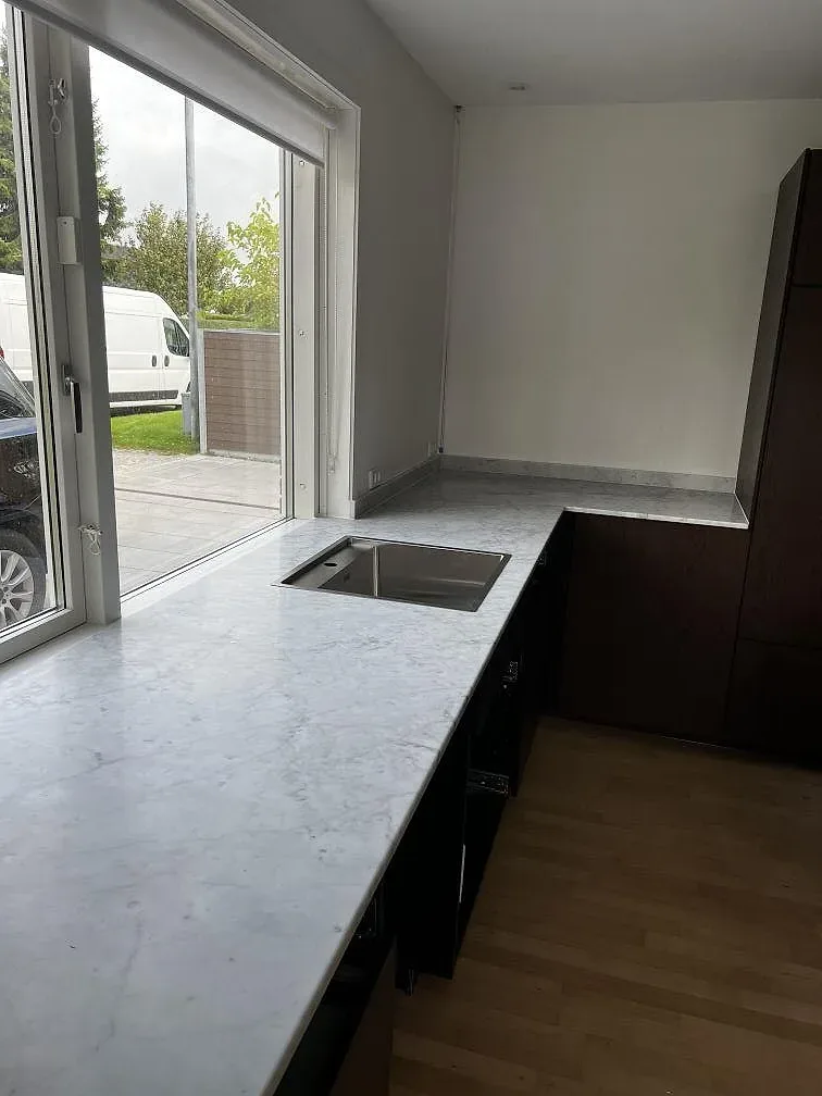 Matt köksbänkskiva i Carrara C marmor i L-form med rostfri diskho, svart häll och trägolv i ett kök i Greve.
