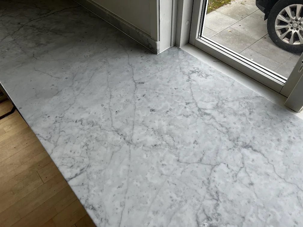 Ovanvy av matt Carrara C köksbänkskiva vid glasdörrar, med vit botten och grå marmorådring i Greve.