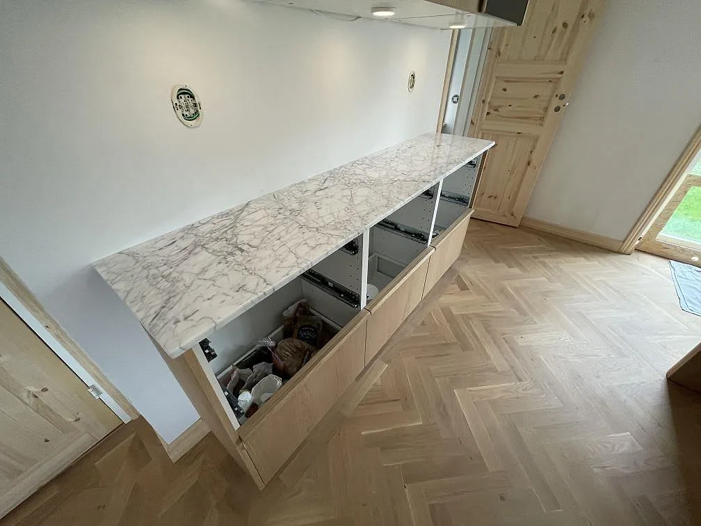 Lång bänkrad i polerad Carrara C marmor med tydlig grå ådring ovanför öppna ljusa köksstommar i Hägersten.