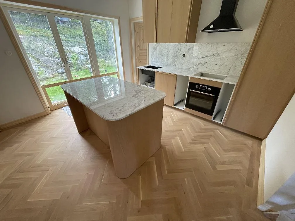 Köksbänkskiva och köksö i polerad Carrara C marmor med grå ådring, väggbeklädnad i samma sten och öppningar för häll och diskho i ett kök i Hägersten.