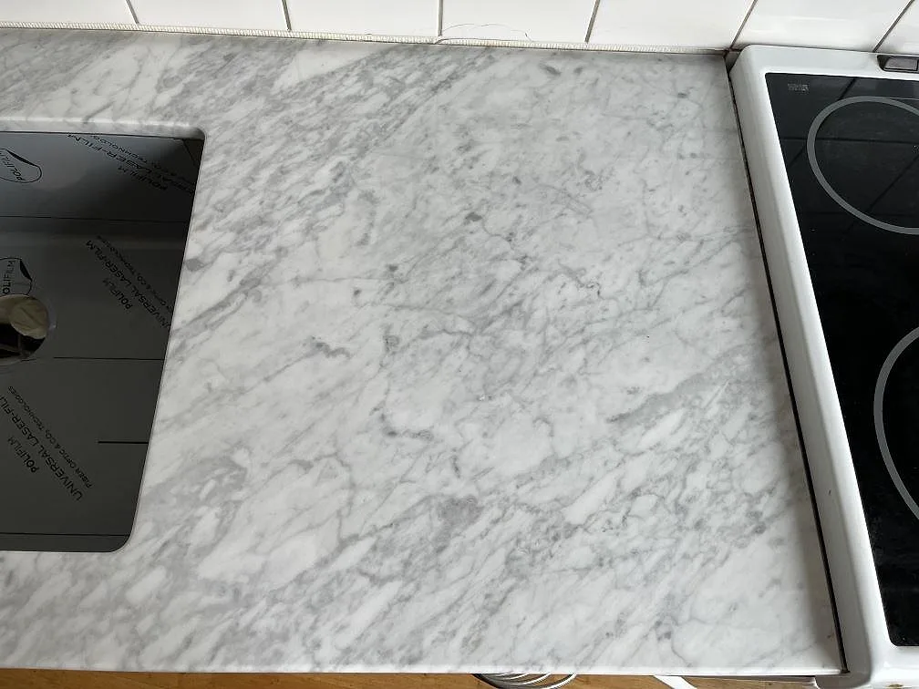 Köksbänkskiva i vit Carrara-marmor med grå ådring, polerad, monterad bredvid en svart glaskeramikhäll och vit kakelvägg i ett kök i Helsingborg.