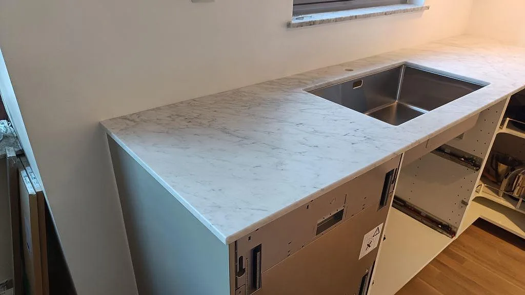 Köksbänkskiva i 20 mm matt Carrara C marmor med synlig rostfri diskho över öppna stommar i ett kök i Helsingborg.