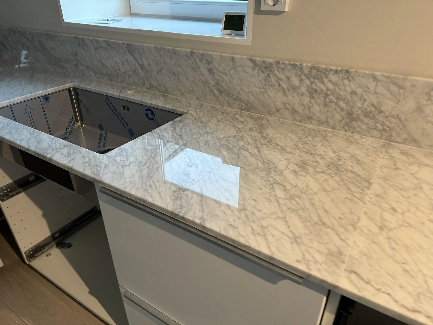 Lång bänkyta i polerad Carrara C-marmor med uppvikt stänkkant och öppna vita köksstommar i Höganäs.