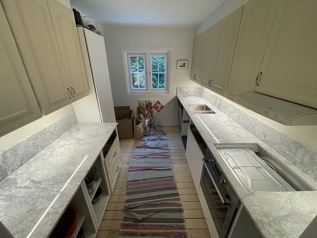 Köksbänkskiva i Carrara C-marmor, polerad, längs två väggar i ett smalt parallellkök i Huddinge, med uppvikt stänkkant, underlimmad rostfri diskho vid fönstret och urtagning för häll.