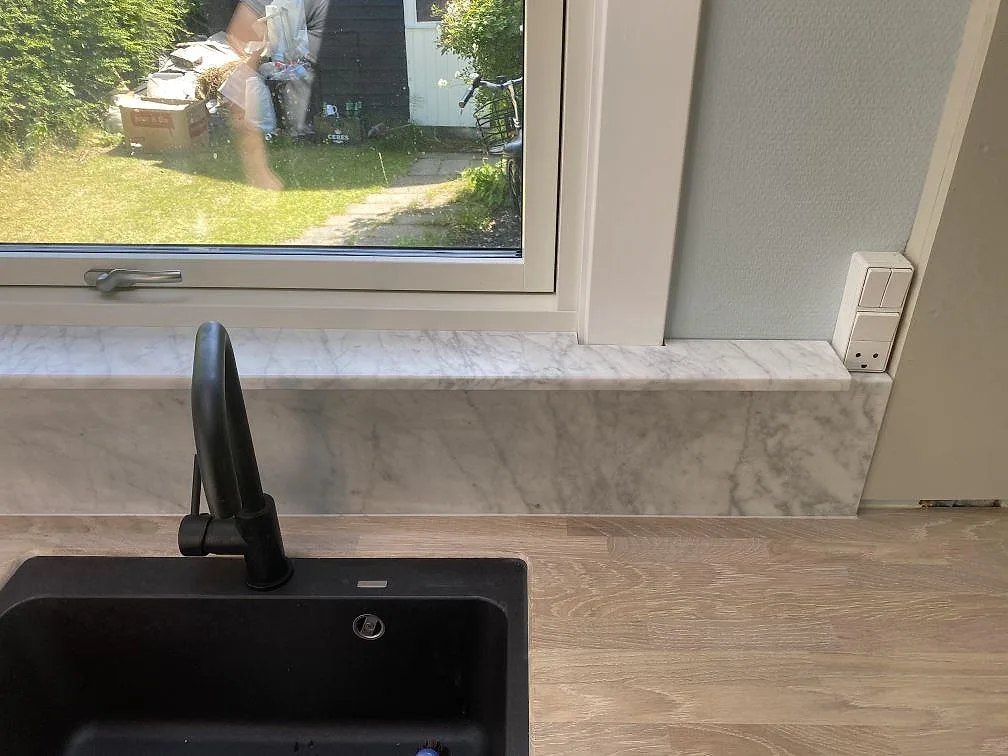 Närbild på fönsterbänk i vit Carrara C-marmor med svart blandare och underliggande kompositdiskho i ett kök i Jægerspris.