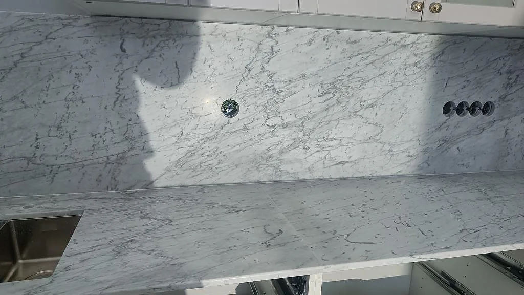Lång köksbänkskiva och hög väggskiva i Carrara C marmor, Matt, med eluttag under montering i Klagstorp.