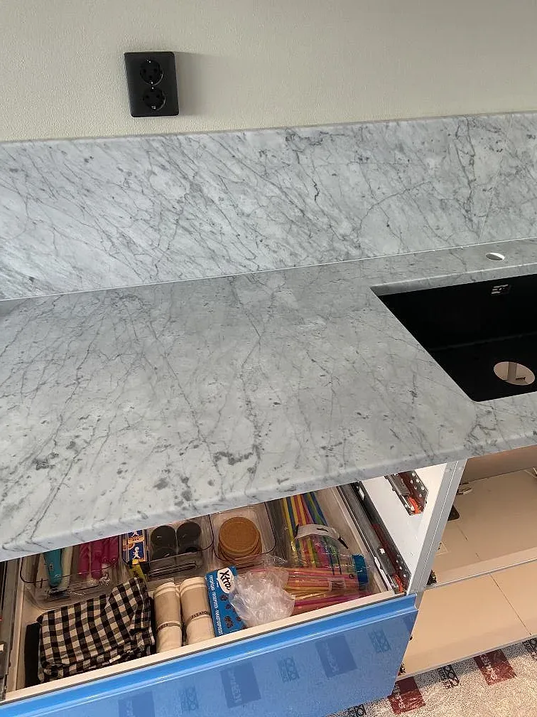 Köksö i Carrara C marmor med uttagad häll och öppna lådor i ett kök under montering i Landskrona.