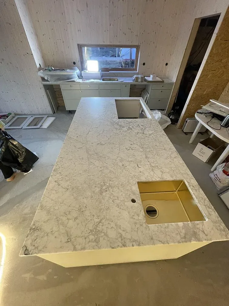 Köksbänkskiva i vit Carrara C-marmor med grå ådring, matt finish, monterad som köksö med underlimmad diskho i ett kök under montering i Leksand.