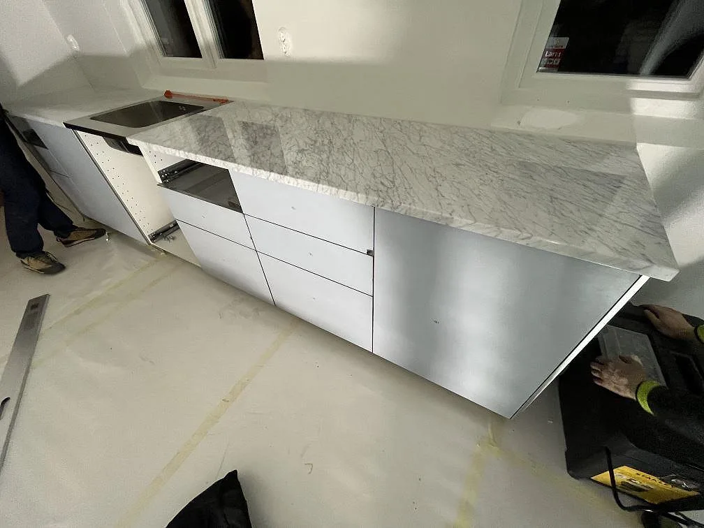 Lång polerad bänkskiva i Carrara C-marmor under fönster med synlig diskhozon och kraftig ådring i ett kök i Lidingö.