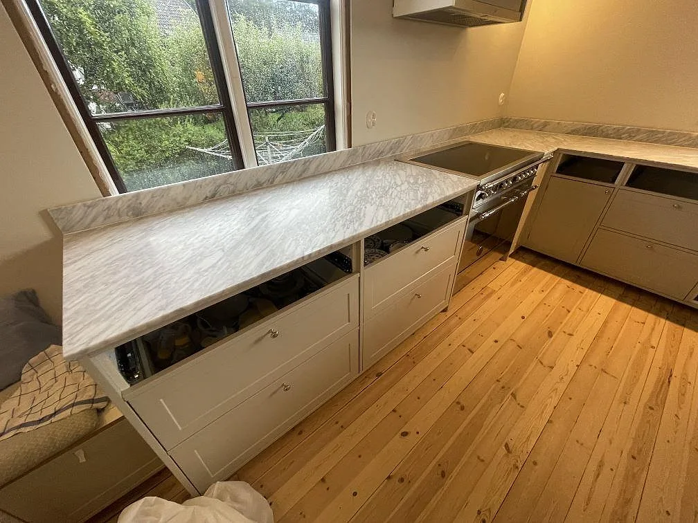 Köksbänkskiva i Carrara C marmor, matt, med uppvikt stänkkant och spis i ett kök i Linköping.