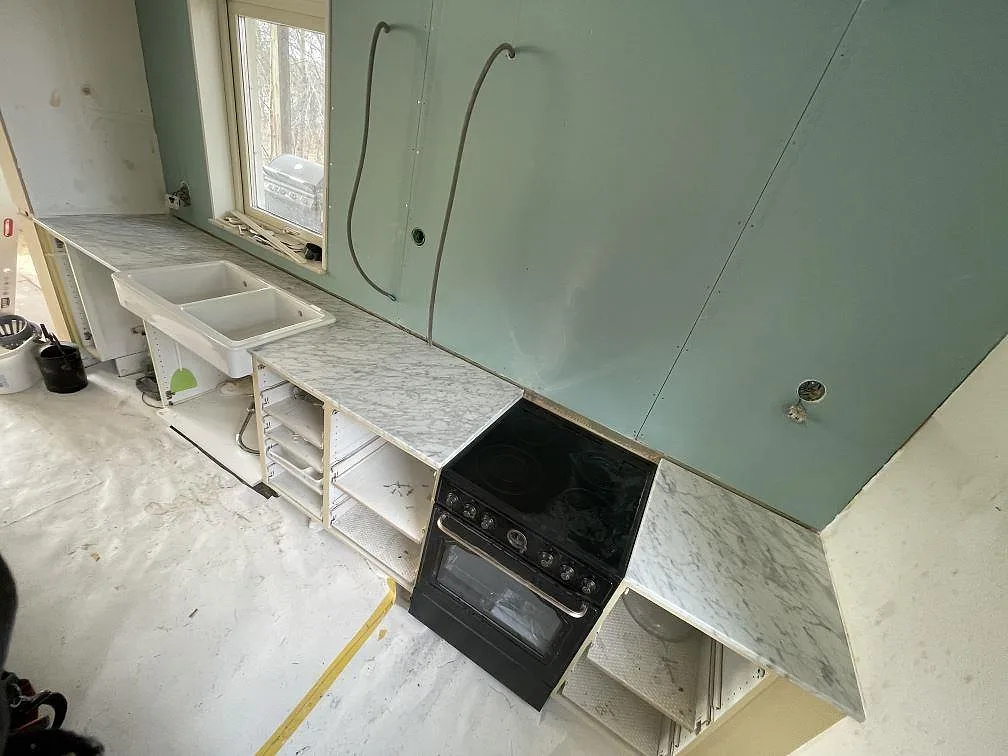 Köksbänkskiva i vit Carrara C-marmor, 20 mm matt, som löper runt hörnet längs två väggar med vit keramisk dubbeldiskho och plats för fristående spis i ett kök på Ljusterö.