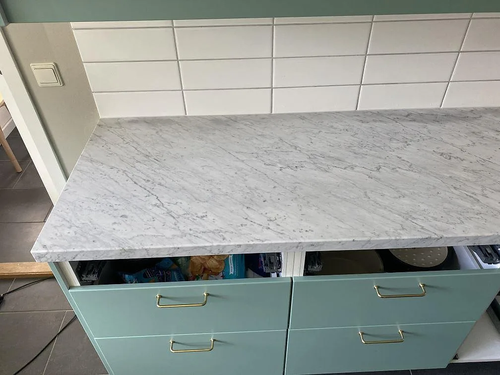 Köksbänkskiva i vit Carrara C marmor, matt yta, med tydlig grå ådring över gröna lådfronter i ett kök i Löddeköpinge.