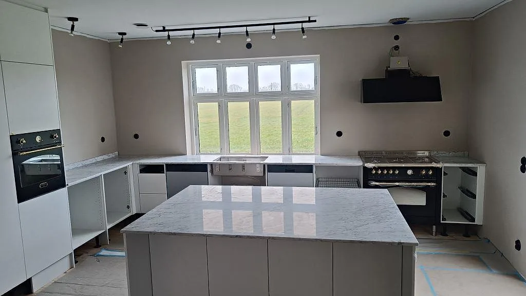 Kök i Lund med köksbänkskiva och köksö i vit Carrara C marmor, fotograferat under montering med spis och fönster i bakgrunden.