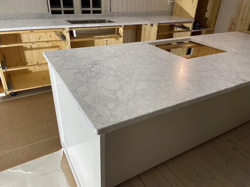 Stor köksö i matt Carrara C marmor med hälluttag framför bänkrad med rostfri diskho i Lycke.