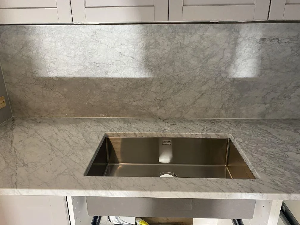 Köksbänkskiva i Carrara C marmor med underlimmad rostfri diskho och hög marmorvägg i grått ådrad marmor i ett kök i Malmö.