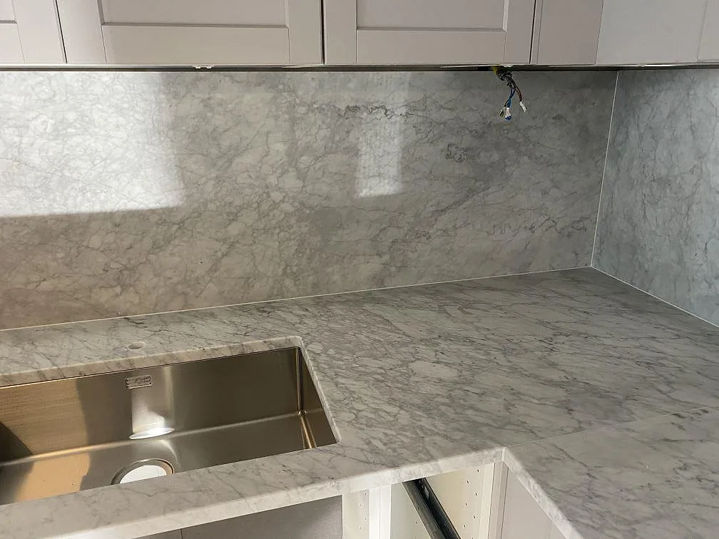 Carrara C marmorbänkskiva i matt finish med underlimmad rostfri diskho i hörnet och uppvikt marmorvägg i ett kök i Malmö.