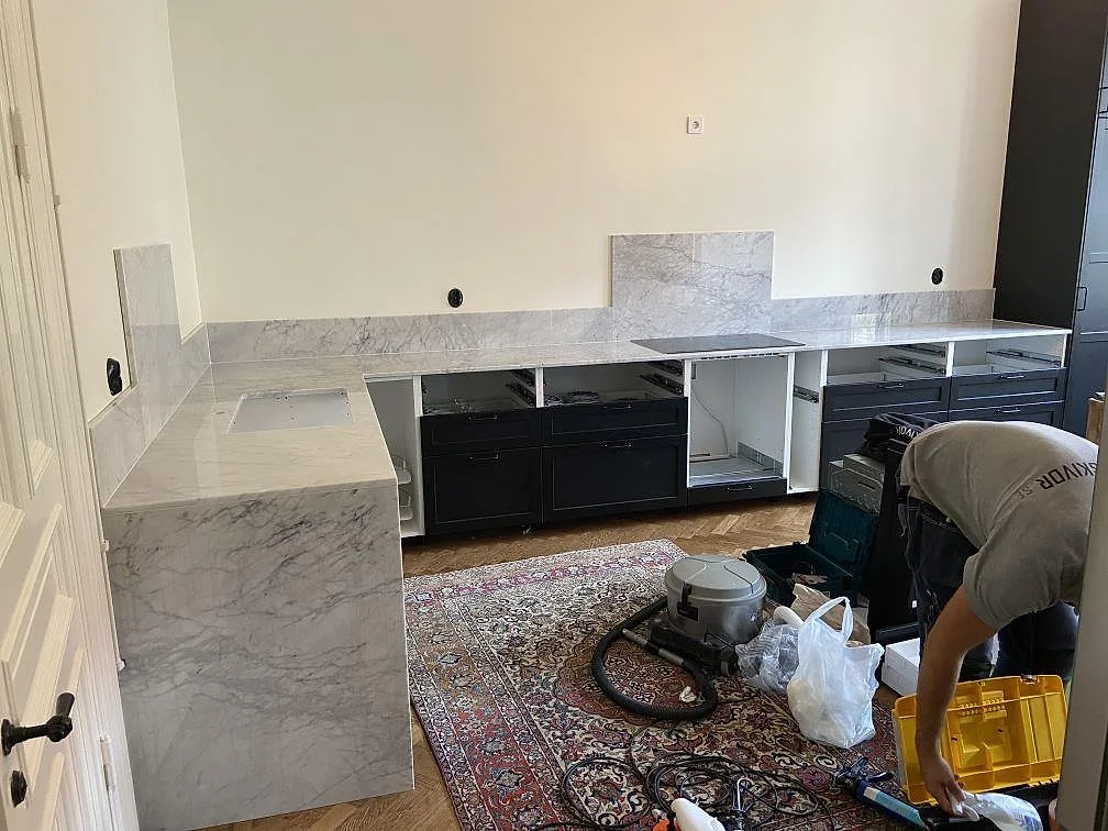 Köksbänkskiva i vit Carrara C marmor med grå ådring i ett vinklat kök i Malmö, med fristående köksö, infälld induktionshäll och urtagning för diskho under montering.