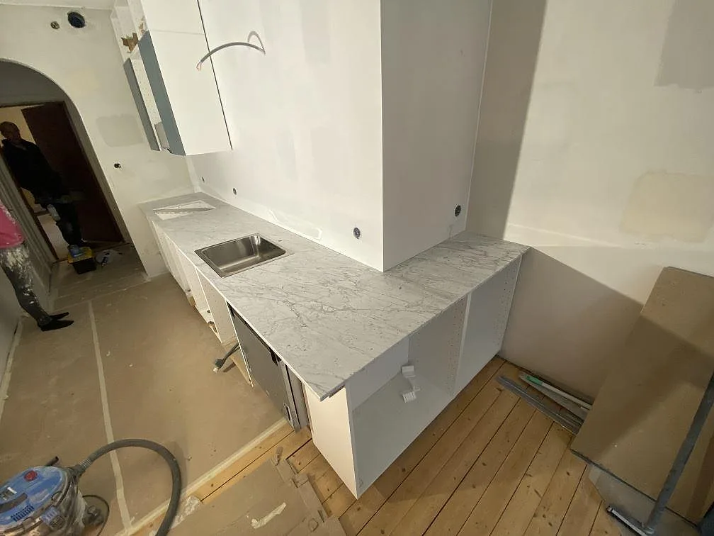 Köksbänkskiva i matt Carrara C marmor med grå ådring i ett kök under montering i Malmö, med rostfri diskho och vita köksstommar.