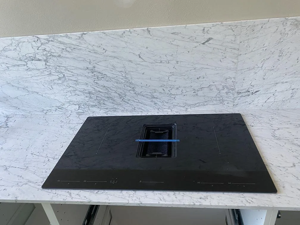 Svart induktionshäll med integrerad fläkt infälld i en vit Carrara C marmorbänkskiva i ett kök i Malmö.
