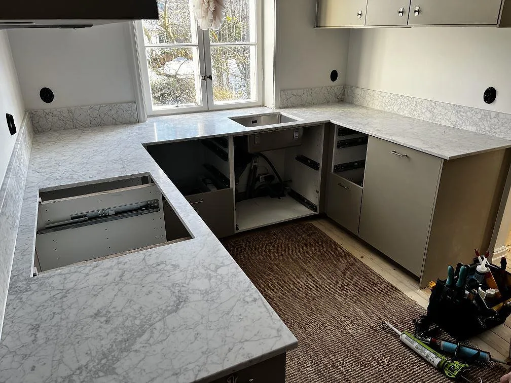 U-formad köksbänkskiva i matt Carrara C marmor, 20 mm, med stänkkant och underlimmad rostfri dubbelho i ett kök i Malmö.