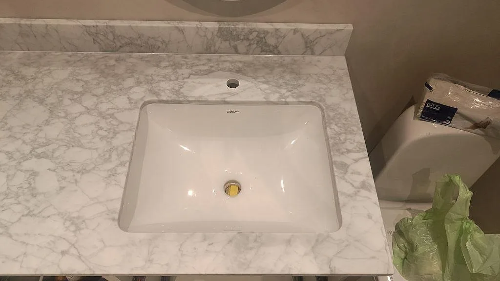 Ovanifrån syns badrumsbänkskivan i matt Carrara C marmor med vitt underlimmat Duravit-tvättställ och blandarhål i Malmö.