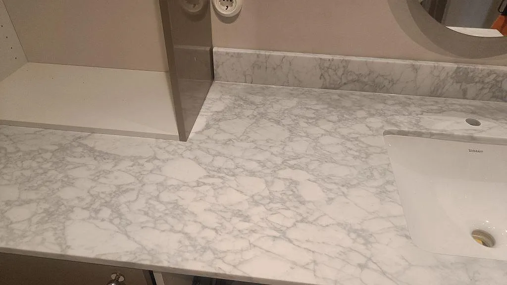 Närbild av matt Carrara C marmorskiva med grå ådring, uppvikt stänkkant och del av vitt tvättställ i ett badrum i Malmö.