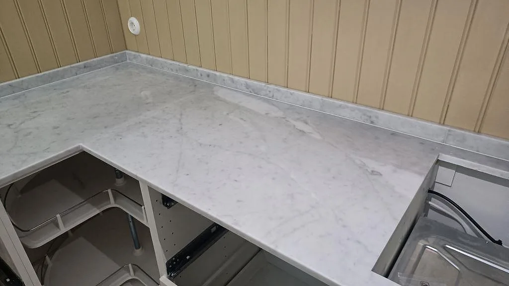 L-formad bänkskiva i polerad Carrara C marmor med låg stenkant längs träpanel i Malmö.