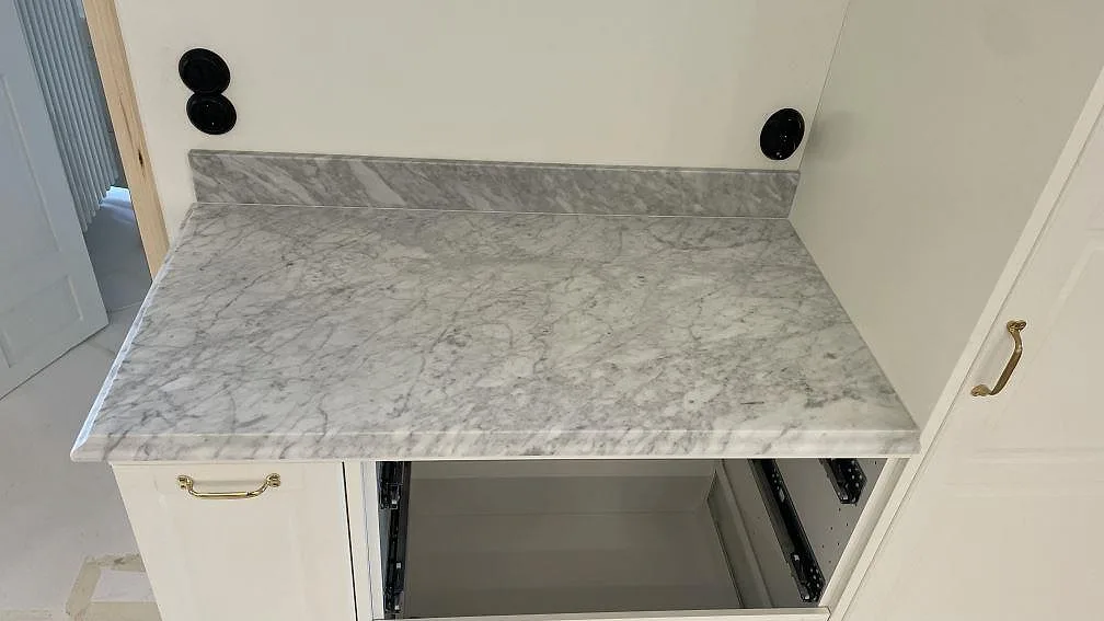 Sidobänk i matt Carrara C marmor med uppvikt stänkkant och öppen köksstomme under montering i Malmö.