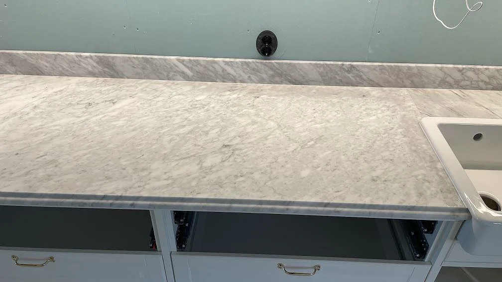 Lång köksbänkskiva i matt Carrara C marmor med ådrad yta, stänkkant och vit dubbelho vid kanten i Malmö.