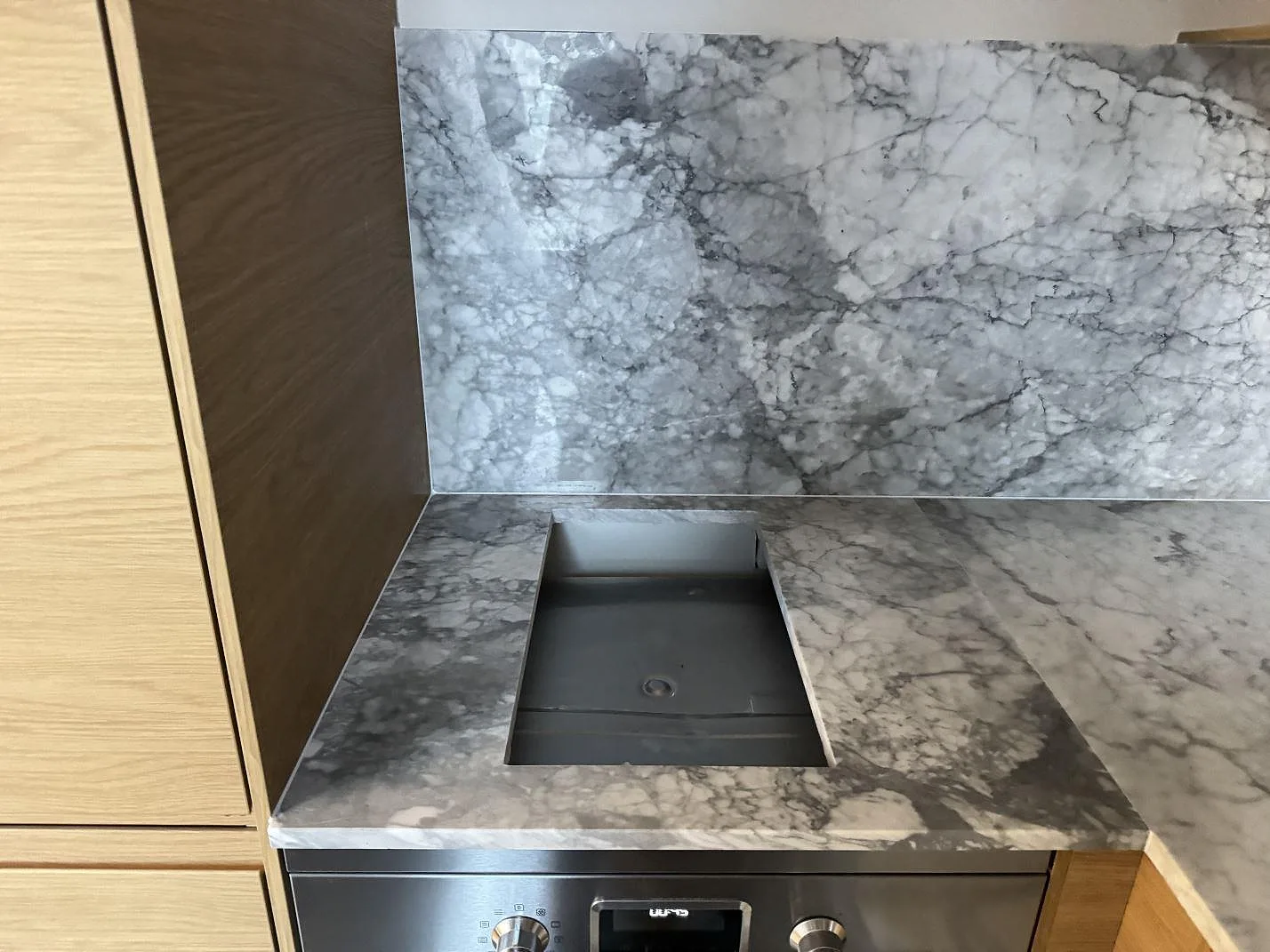 Köksbänkskiva i Carrara C, polerad naturmarmor med grafitgrå ådring, i ett L-format kök i Malmö med underlimmad rostfri diskho och urtag för häll under montering.