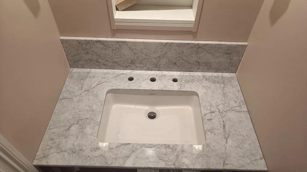 Närbild ovanifrån av polerad marmorbänkskiva i Carrara C med synlig ådring, tvättställ och tre blandarhål i Mariestad.