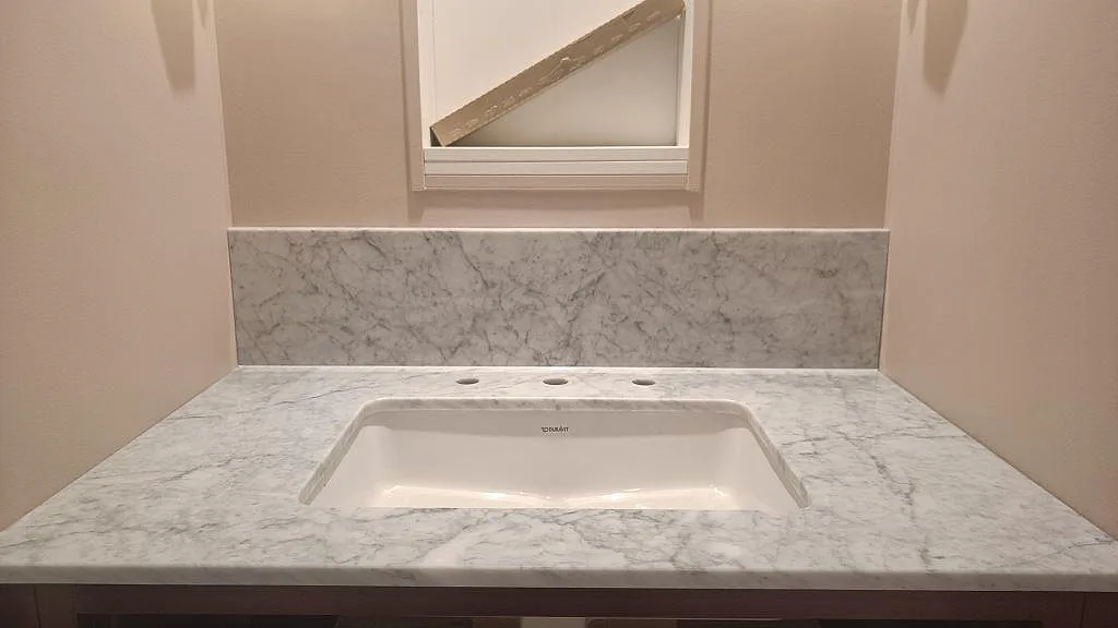 Frontal bild av polerad Carrara C marmor som badrumsbänkskiva med tvättställ och stänkkant under fönster i Mariestad.