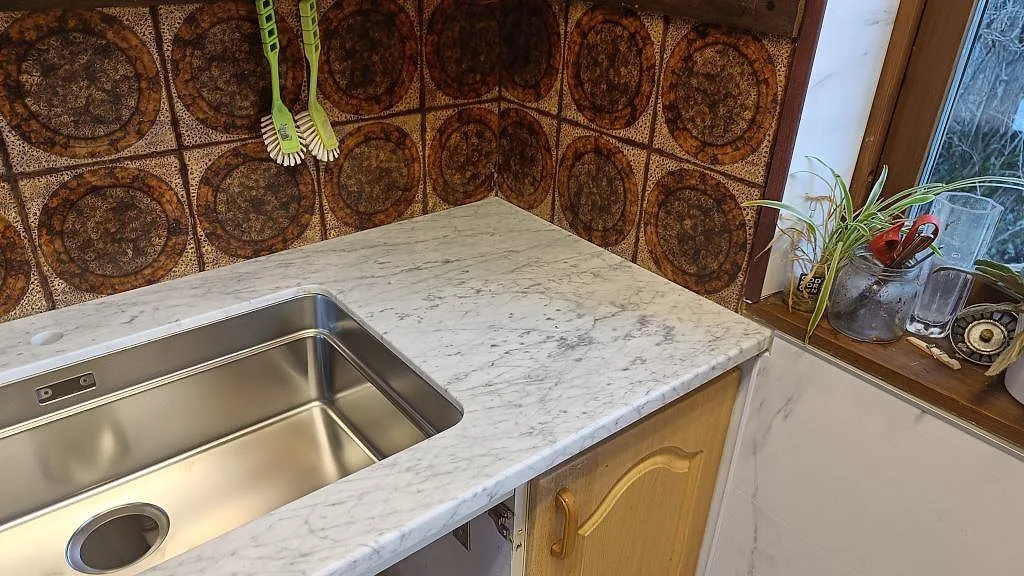 Detalj av Carrara C vid diskho och fönsterhörn, polerad marmorbänkskiva med grå ådring i ett kök i Mellbystrand.