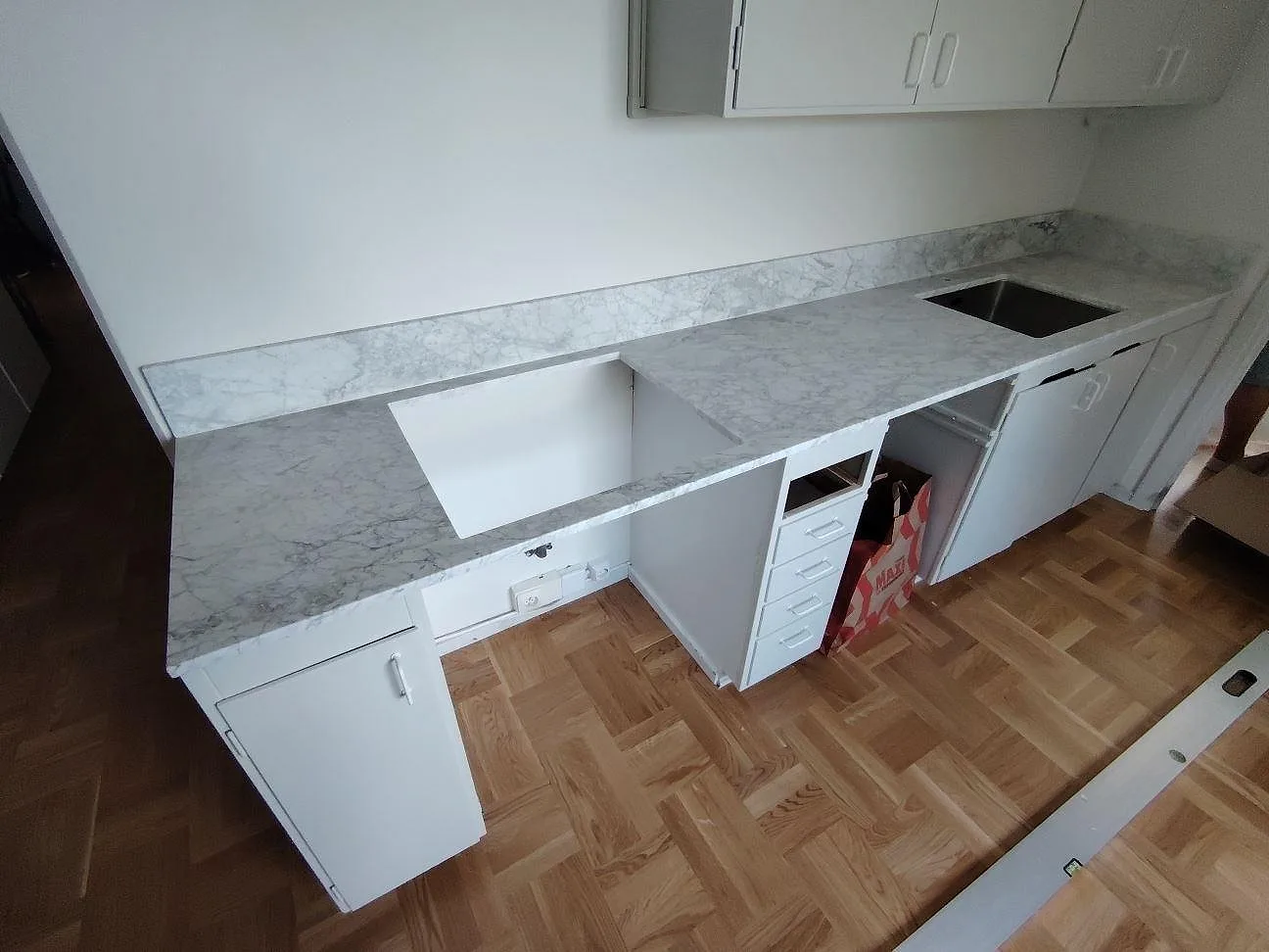 Köksbänkskiva i matt Carrara C-marmor med L-formad arbetsyta, stänkkant och öppningar i ett kök i Nacka.