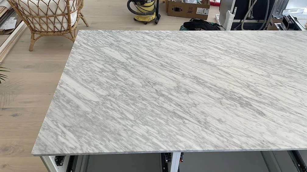 Stor köksö i matt Carrara C marmor, 20 mm, med grå ådring och öppna köksstommar under montering i Nättraby.