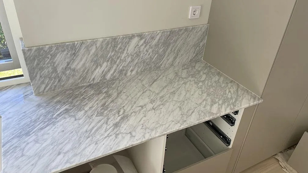 Sidobänk i matt Carrara C marmor med låg stänkkant mot högskåp i ett kök under montering i Nättraby.