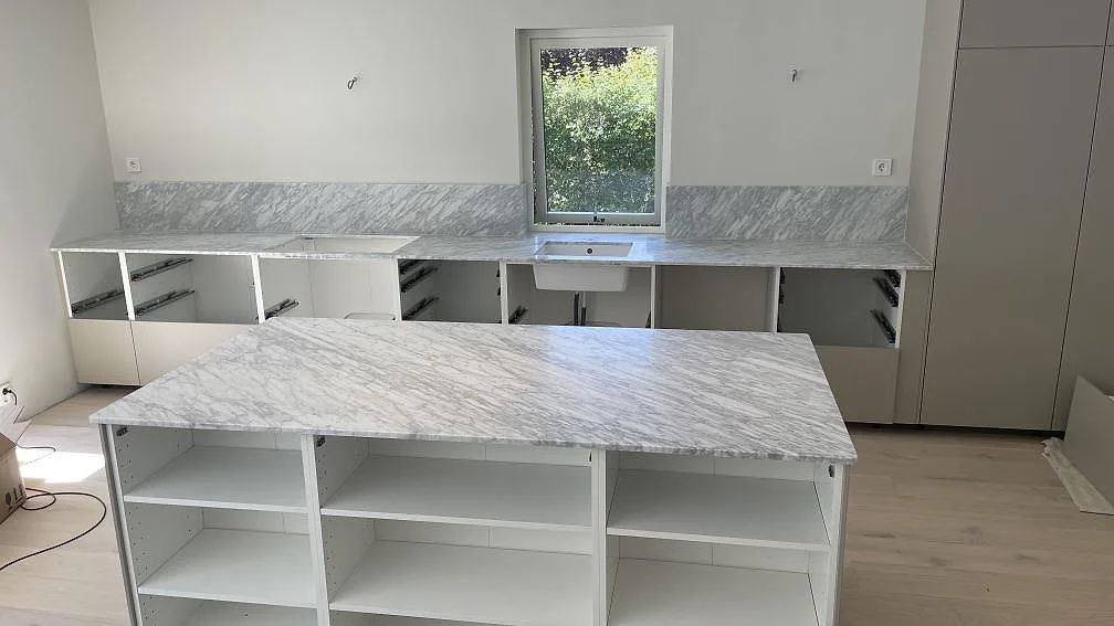 Köksbänkskiva och stor köksö i matt Carrara C marmor, 20 mm, med låg stänkkant och diskplats under fönstret i Nättraby.