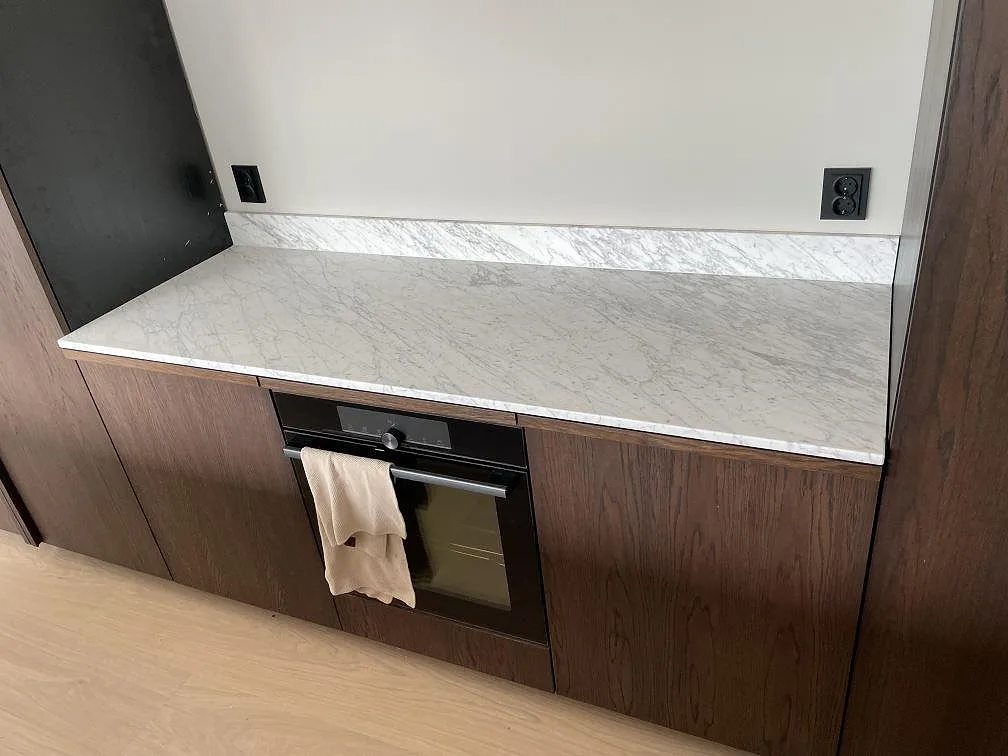 Väggbänk i matt Carrara C marmor, 20 mm, med låg stänkkant och grå ådring över mörka köksskåp i Norrtälje.