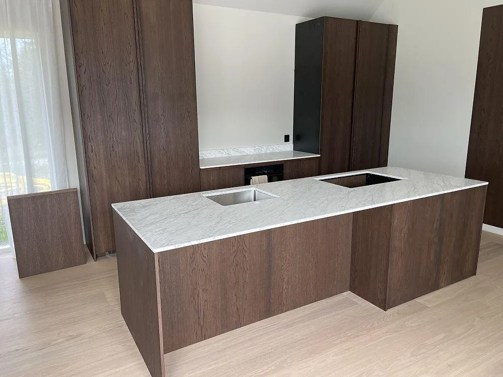 Köksbänkskiva i matt Carrara C marmor, 20 mm, på mörk träklädd köksö med synliga urtag i Norrtälje.