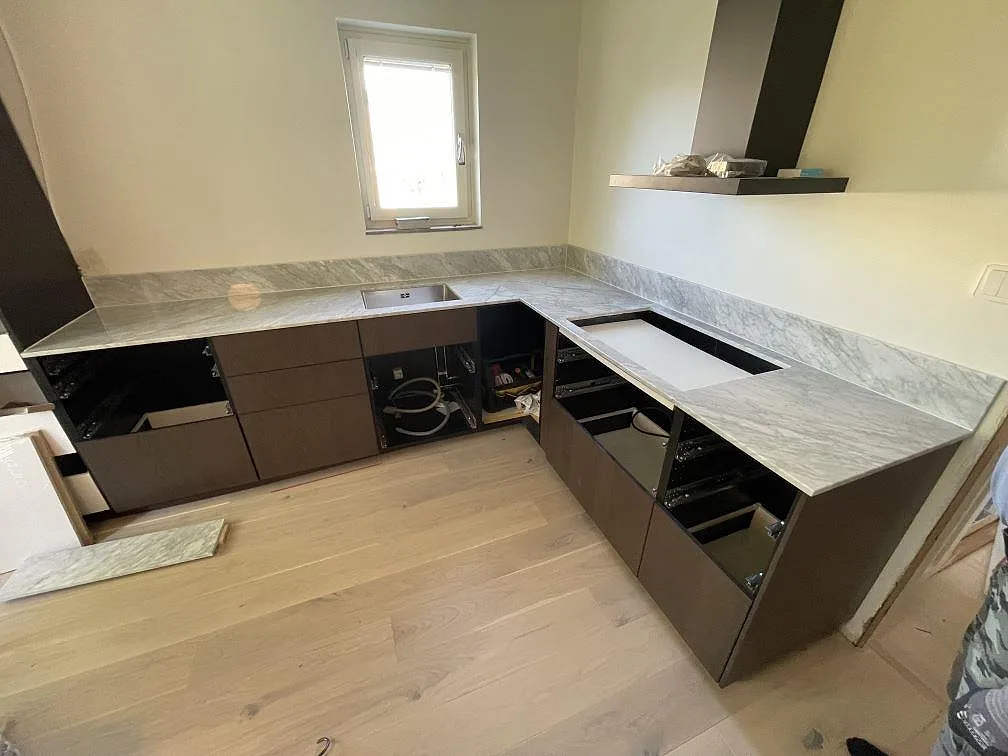Köksbänkskiva i polerad Carrara C-marmor med diskho, stänkkant och ljus grå ådring i ett kök under montering i Norrtälje.