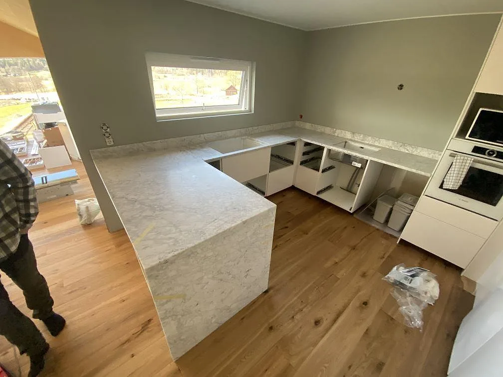 Köksbänkskiva i vit Carrara C marmor, matt yta, i ett U-format kök under montering med fönster och ekgolv i Nygård.
