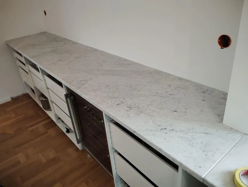 Lång köksbänkskiva i matt Carrara C marmor med synlig grå ådring i ett kök under montering i Örebro.